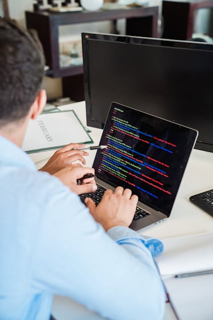 A person wearing a light blue shirt is seated at a desk, engaged in a home relocation project involving packing and moving logistics. They are using a laptop displaying a screen filled with colorful l