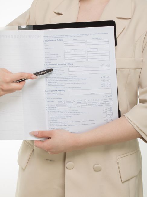 A professional woman with long black hair, dressed in a grey checkered vest and light-colored top, is holding a clipboard with a home insurance policy and a pen, engaging in a conversation with a cust