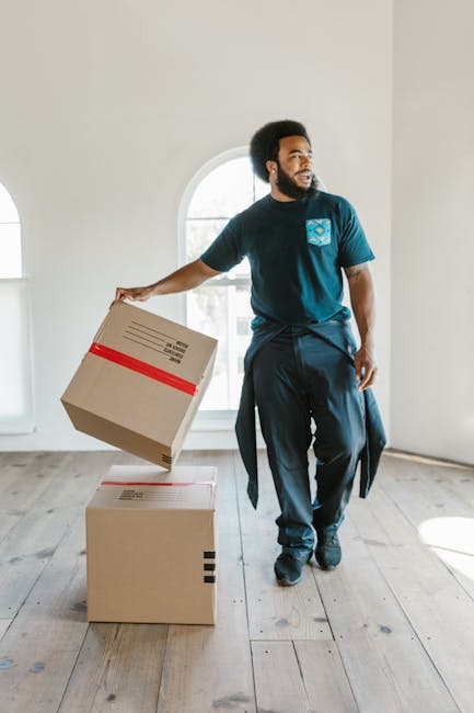 Two movers from Removal Companies Brixton are engaged in a home relocation task, loading a large, dark green wrapped piece of furniture into the open cargo area of a white panel van. The van is parked