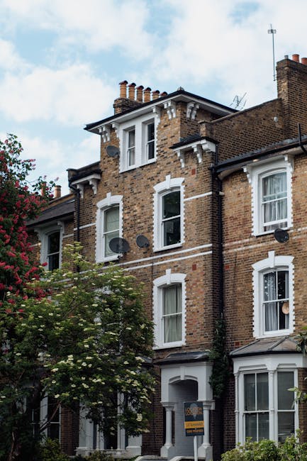 A row of traditional terraced houses on Coldharbour Lane Brixton, with brick facades and bay windows, some open with curtains visible. The front gardens are enclosed by black metal fences, with a few 
